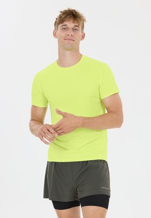 Ung mand iført en lys gul kortærmet skjorte og mørkegrå lagdelte atletikshorts, stående mod en hvid baggrund.