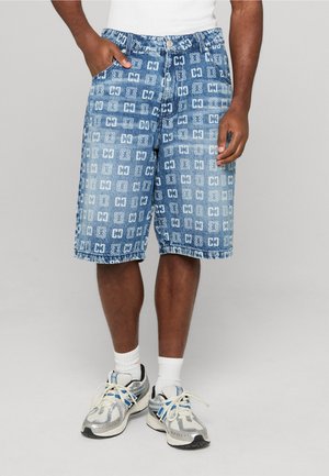 Man draagt blauwe spijkershorts met patroon, witte sokken, zilveren sneakers en een wit mouwloos topje, staat met een hand in de zak.