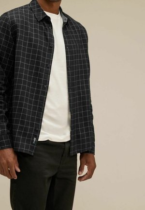 Uomo che indossa una camicia nera a quadri aperta a maniche lunghe sopra una maglietta bianca e pantaloni neri su uno sfondo beige uniforme.