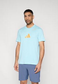 Ανοιχτό μπλε κοντομάνικο με στρογγυλή λαιμόκοψη, που διαθέτει πορτοκαλί ριγωτό λογότυπο στο στήθος. Φοράται με μπλε σορτς. Λεία υφασμάτινη υφή.