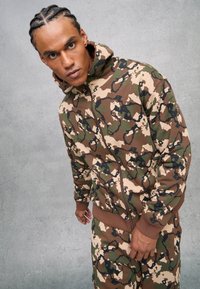 Uomo con i capelli intrecciati che si inclina in avanti, indossando un felpa con zip camouflage marrone, verde e beige e pantaloni abbinati su uno sfondo grigio.