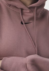 Pehmeästä kankaasta valmistettu vaaleanpunainen huppari, jossa on musta Nike-swoosh-logo, kiristysnyörit ja rento istuvuus resoreilla varustetuissa hihoissa.