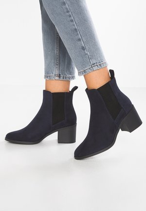 Enkelhoge laarzen in donkerblauw met blokhakken, gedragen bij lichtblauwe cropped jeans op een witte vloer.