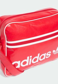 Borsa rossa Adidas realizzata in materiale sintetico lucido, con accenti bianchi, una zip frontale e un grande logo su un lato. Forma rettangolare.