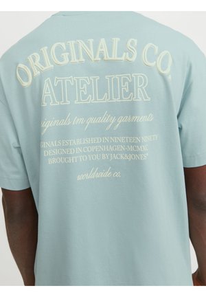T-shirt bleu clair avec un texte crème indiquant "ORIGINALS CO. ATELIER" et une impression plus petite au dos, porté par une personne.