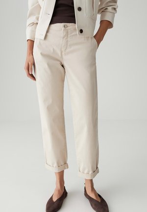 Persona con pantaloni beige risvoltatI, una giacca leggera sopra una maglia scura e scarpe scure senza lacci, in piedi con una mano in tasca.