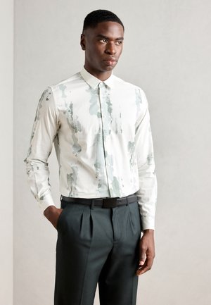 Camicia bianca con motivo astratto verde, design con bottoni, maniche lunghe, abbinata a pantaloni plettati scuri e una cintura nera.
