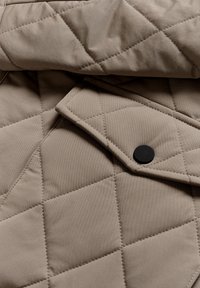 Tejido acolchado beige con patrón de diamantes, que presenta un cuello doblado y un detalle de botón a presión negro. Textura suave con acabado liso.