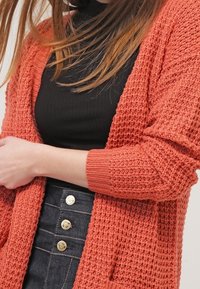 Cardigan a maglia texturizzata di color ruggine, con apertura frontale, maniche lunghe e tasche, indossato sopra una camicia nera aderente e una gonna di jeans con bottoni.