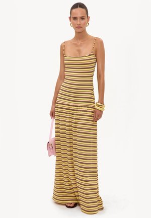 Femme portant une robe longue sans manches rayée jaune et marron, tenant un sac à main rose, avec des boucles d'oreilles créoles dorées et des bracelets.