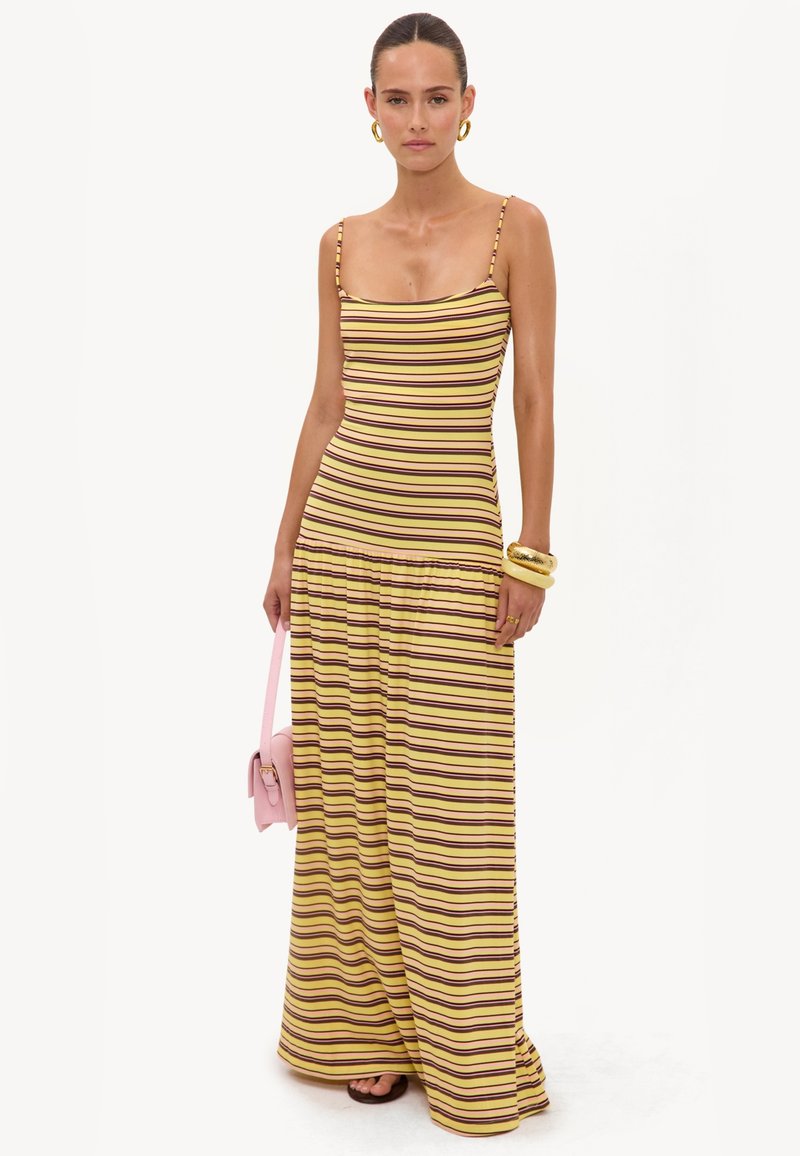 Femme portant une robe longue sans manches rayée jaune et marron, tenant un sac à main rose, avec des boucles d'oreilles créoles dorées et des bracelets.