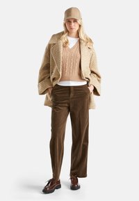 Wełniany płaszcz w hektarowatym wzorze w kolorze tan, dzianinowy sweter w jasnym brązie, biały t-shirt, brązowe spodnie kordowe, brązowe buty, beżowa czapka. Widoczne teksturowane materiały.