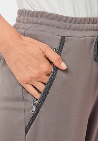 Graue Jogginghose mit verstellbarem Bund, ausgestattet mit einer silbernen Reißverschlusstasche mit dunklem Besatz und einem glatten, strukturierten Stoff.