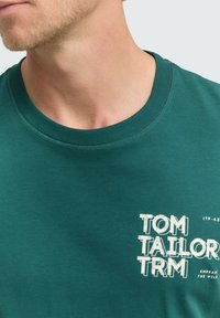 Camiseta de algodón verde azulado con cuello redondo, mangas cortas y un estampado gráfico en el pecho que incluye el nombre de la marca y el eslogan en blanco.