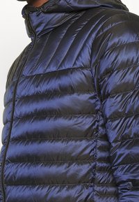 Marinblå pufferjacka med horisontell sömnad, ståkrage och dragkedjestängning. Texturerad satinliknande finish med en slät yta.