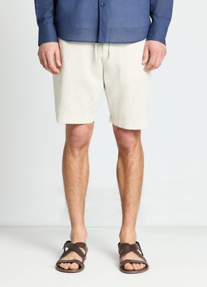 Uomo con pantaloncini color crema, sandali marrone scuro e camicia blu a maniche lunghe, in piedi su un pavimento e uno sfondo bianchi.