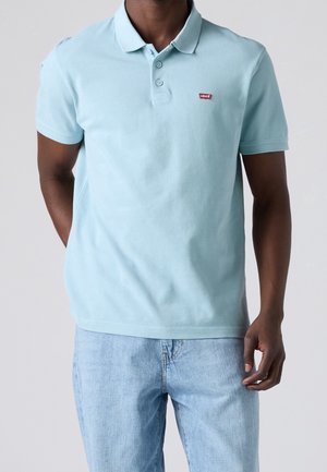 Mand iført lyseblå Levi's poloshirt og lyseblå jeans står med afslappede hænder.