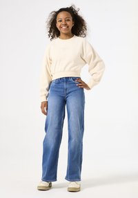 Sudadera corta de color crema con mangas anchas, combinada con jeans acampanados de color azul claro y zapatos tipo slip-on beige, de pie sobre un fondo liso.