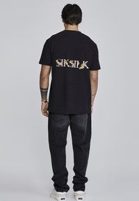 Camiseta de algodón negra con un logo decorativo en dorado y blanco de "SRSILK" en la parte trasera. La persona lleva pantalones oscuros y zapatillas blancas.