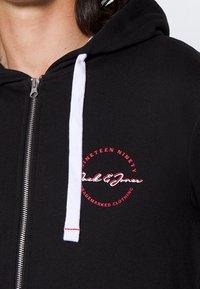 Sudadera negra con cremallera y cordones blancos, y un logo circular rojo con el texto "Jack & Jones" en el pecho.
