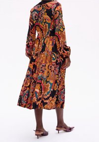 Robe maxi à manches longues, à motifs, dans des tons de marron, noir et couleurs vives, avec une taille cintrée et une coupe ample. Complétée par des chaussures à talons.