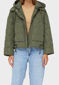 Olivgrüne gesteppte Jacke mit Kapuze, Tunnelzug am Saum und Reißverschluss, getragen über einem beigen Oberteil und kombiniert mit hellblauen Jeans.