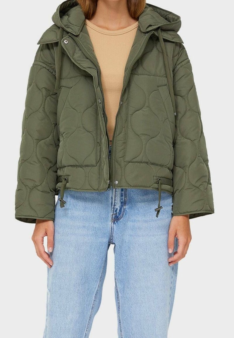 Olivgrüne gesteppte Jacke mit Kapuze, Tunnelzug am Saum und Reißverschluss, getragen über einem beigen Oberteil und kombiniert mit hellblauen Jeans.