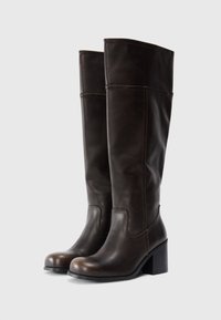 Bottes en cuir marron montantes, avec un bout arrondi, un talon épais et de simples coutures. Texture lisse avec une finition polie.