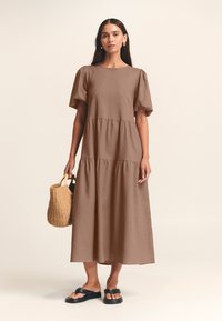 Abito marrone di media lunghezza realizzato in tessuto leggero, con maniche corte a palloncino e gonna a più strati. La modella tiene in mano un cesto intrecciato. Sandali neri.