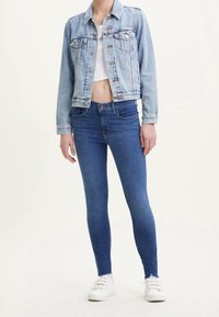 Chaqueta de mezclilla en azul claro con un diseño clásico, dos bolsillos en el pecho y cierres de botón. Jeans skinny azul oscuro con dobladillos deshilachados. Zapatillas blancas.