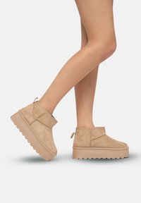 Botas de tobillo de ante beige con una suela de plataforma gruesa, que cuentan con una lengüeta en la parte posterior y acentos en las costuras laterales. Textura suave, diseño casual.