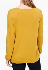 Långärmad senapsgul topp med rundad halsringning, mjukt tyg och texturerad yta, med en avslappnad passform och något rundad fåll.