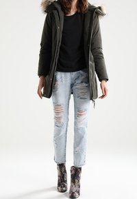 Parka vert foncé avec une capuche bordée de fourrure, associée à un jean bleu clair délavé et à des bottines fleuries à finition brillante.