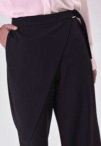 Pantaloni neri a gamba larga con design avvolgente, realizzati in un tessuto morbido, dotati di un laccio laterale e un accento minimalista in argento in vita.