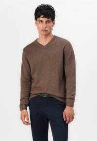 Barna V nyakú pulóver hosszú ujjakkal, texturált anyagból, laza fit, sötét nadrággal és övvel párosítva. Egyszerű háttér.