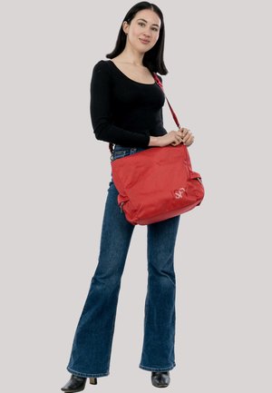 Sieviete ar taisniem tumšiem matiem, tērpusies melnā garroku krekliņā un zilās džinsās, tur lielu sarkanu plecu somu ar divām sānu kabatām.