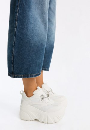 Gambe con jeans blu a gamba larga e sneakers bianche con suola platform e catene decorative argentate sui lacci.