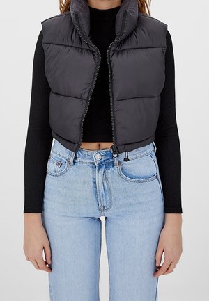 Kvinde iført en sort kort puffervest over en sort langærmet top og lyseblå højtaljede jeans, vist fra nakke til hofter.
