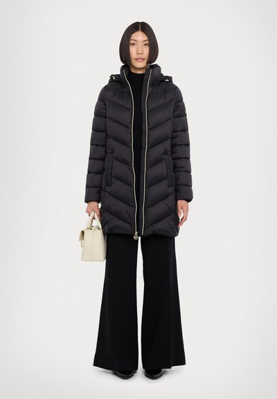 Manteau matelassé noir avec motif en chevrons, fermeture éclair à l'avant et capuche. Dispose de deux poches latérales et de finitions dorées. Porté avec un pantalon noir à jambes larges.