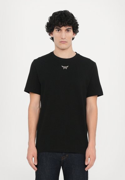 T-shirt en coton noir à manches courtes et col rond, avec un accent logo métallique sur le devant. Porté avec un jean bleu foncé.