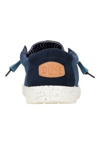 Des chaussures en suède bleu marine à enfiler avec une semelle blanche mouchetée, dotées d'une étiquette en cuir beige embossée avec "HEY DUDE" sur le talon.