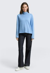 Helles blaues Rollkragensweatshirt aus weichem Strick, mit langen Ärmeln und lockerer Passform, kombiniert mit dunklen, ausgestellt geschnittenen Jeans und weißen Sneakers.