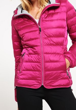 Femme portant une veste matelassée rose vif avec des poches zippées et un pantalon noir, debout avec une main dans la poche.