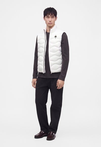 Joven con un chaleco acolchado blanco sin mangas sobre una camisa negra de manga larga, pantalones negros y zapatos marrón oscuro, de pie sobre un fondo sencillo.