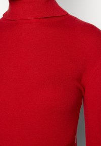 Pull à col montant rouge en tissu tricoté côtelé doux. Il présente une silhouette ajustée et une texture lisse sans motifs ni embellissements visibles.