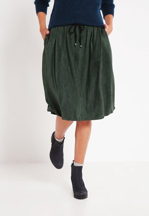Woman wears dark green knee-length skirt with drawstring waist, navy ankle boots, and navy sweater, hands in pockets, walking forward.