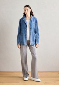 Světlomodrá džínová bunda s klopovým límcem, dvěma předními kapsami a zapínáním na knoflíky, nošená přes bílou košili a šedé kalhoty.