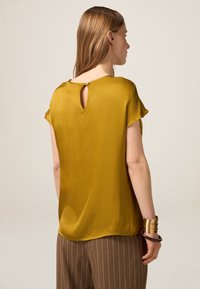 Kurzärmelige Senfgelbe Bluse mit einer Schlüssel-Aussparung am Rücken, glatter Textur und abgerundetem Saum. Kombiniert mit braunen gestreiften Hosen mit hohem Bund.
