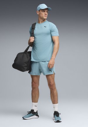 Hombre vistiendo conjunto deportivo Puma azul claro y gorra, sosteniendo una bolsa de gimnasio negra, de pie contra un fondo gris liso.