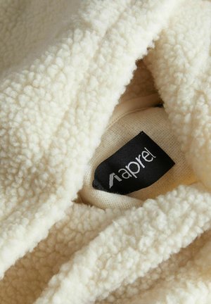 Közeli kép egy puha, krémszínű, texturált polárszövetről, amelyben részben látható egy fekete-fehér címkés címke.